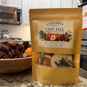 May include: A tan paper pouch containing a simmer pot blend labeled Chavarría Home Co. Cozy Fall. The label features illustrations of apple, orange, and cinnamon. A clear window reveals dried apple slices, orange, and cloves inside the bag.
