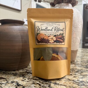 May include: A tan stand-up pouch containing a simmer pot blend labeled Woodland Retreat by Chavarria Home Co. The label features illustrations of orange slices, cinnamon sticks, and star anise. A clear window at the bottom reveals dried bay leaves and orange slices inside the bag.