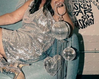 May include: A silver sequin mini dress, a white bridal veil, and metallic silver knee-high boots. The ensemble includes a circular disco ball handbag with a heart-shaped charm and long dangling silver chains, held by a person.