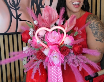 May include: A vibrant floral bouquet featuring pink and red artificial flowers, feathery plumes, and a central glowing neon heart shape. The arrangement is accented with silver glitter spheres and long, trailing pink ribbons with white polka dots.
