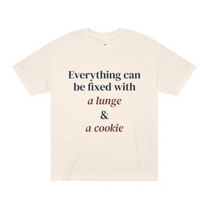 Op de afbeelding: Een crèmekleurig t-shirt met korte mouwen en de donkerblauwe en bordeauxrode tekst: Everything can be fixed with a lunge & a cookie. Het t-shirt heeft een ronde hals en een standaard pasvorm, met de bedrukte tekst gecentreerd op de borst.