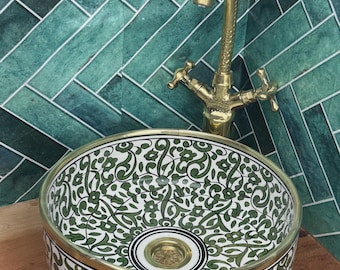 May include: A round ceramic vessel sink featuring a white base with intricate green floral and vine patterns, finished with a brass rim and a matching brass drain. A tall, curved brass faucet with two cross-handle knobs stands behind the sink.