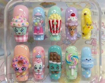 May include: A set of ten press-on nails in a clear plastic case, each featuring a unique 3D charm inspired by food and candy characters, including a cupcake, gumball machine, popcorn, ice cream, banana, donut, milk carton, chocolate bar, lollipop, and macaron.