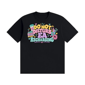 May include: A black t-shirt featuring a colourful graphic with the text DO NOT DISTURB EA RECHARGING. The design includes illustrations of a coffee cup, a stack of books labelled SUPPORT, ENCOURAGE, INCLUDE, BELIEVE, a pencil holder, and a scrunchie.