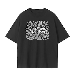 May include: A black short-sleeved t-shirt featuring a white graphic print on the chest. The design includes the text "EDUCATIONAL ASSISTANT" surrounded by line-art illustrations of a globe, books, a pencil, a backpack, an apple, and hearts.