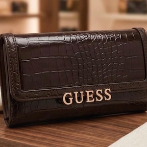 Peut inclure: Un portefeuille rectangulaire marron foncé avec une texture brillante gaufrée façon crocodile. Le rabat avant présente le mot GUESS en lettres métalliques dorées centrées près du bord inférieur. Le portefeuille a une structure rigide.