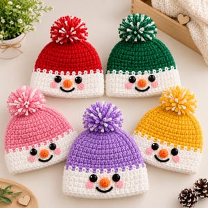 May include: Five crocheted beanies featuring snowman faces with black button eyes, orange noses, and pink cheeks. Each hat has a white brim and a matching colored pom-pom on top in red, green, pink, purple, and yellow yarn colors.