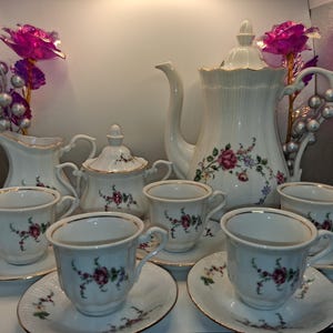 Puede incluir: Un juego de té de porcelana blanca que incluye una tetera alta con tapa, una jarra de leche, un azucarero con tapa y cuatro tazas con sus platos a juego. Cada pieza está decorada con un delicado patrón floral de rosas rosas y hojas verdes.