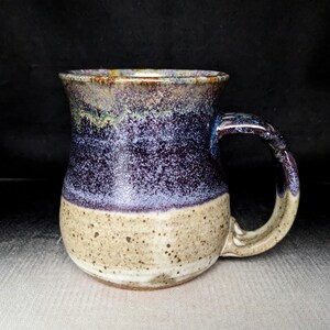 May include: Handmade ceramic mug with a speckled beige base, transitioning to a deep purple and blue speckled body, and a dark handle. The rim features a blend of brown and gold tones. This unique mug is perfect for coffee or tea.
