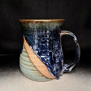 May include: Handmade ceramic mug with a unique design. The mug features a dark blue and white marbled glaze, with a diagonal band of tan and light blue stripes. The rim is a darker brown. The mug has a large handle.