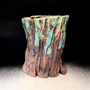 May include: A cylindrical ceramic vessel with a textured, bark-like exterior in shades of dark brown and reddish-brown. The top rim features a decorative glaze in vibrant turquoise and yellow that drips down the sides, mimicking natural wood grain.