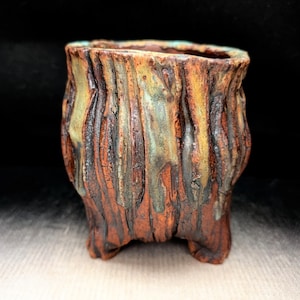 May include: A small, hand-built ceramic pot with a textured, bark-like surface. The exterior features a deep reddish-brown clay base with vertical ridges and streaks of mottled green and yellow glaze dripping down the sides.
