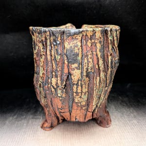 May include: A cylindrical ceramic vessel with a textured, bark-like exterior featuring a mottled color palette of deep brown, burnt orange, and muted green. The pot stands on three small, integrated feet and has a rugged, uneven rim.