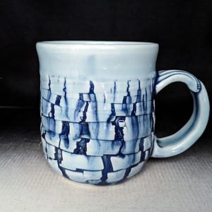 May include: A ceramic mug with a light blue glaze and a textured, brick-like pattern in shades of blue. The handle is curved and matches the mug's color. The mug is designed for hot beverages.