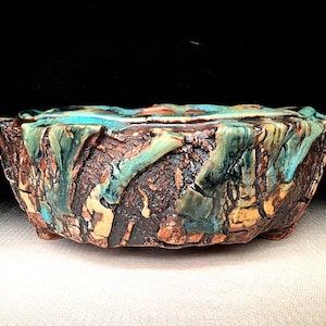 May include: A round ceramic bonsai pot with a textured, bark-like exterior in shades of dark brown and tan. The rim features a vibrant, glossy turquoise glaze that drips down the sides, contrasting with the rough, earthy base and small feet.