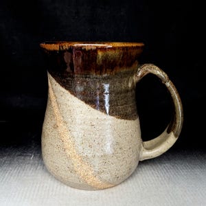 May include: A handmade ceramic mug with a rounded body and a large handle. The mug features a diagonal color block design, with a light beige lower half and a dark brown upper half. The rim is a darker brown. The mug is suitable for hot beverages.