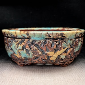 May include: A ceramic bonsai pot with a textured, rugged exterior featuring a mix of deep brown, tan, and turquoise crackle glaze. The bowl-shaped vessel has a wide, rounded rim and sits on four small, integrated feet, showcasing a weathered appearance.