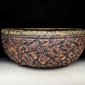 May include: A handcrafted, oval-shaped bonsai pot. The exterior features a textured, bark-like design in shades of brown and orange. The rim is accented with a green and tan trim. The pot sits on a white surface against a black background.