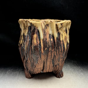 May include: A small, footed ceramic vessel with a textured, bark-like brown exterior and a contrasting cream-colored glaze that drips down from the uneven, scalloped rim. The surface features deep vertical grooves and a rough, organic finish.