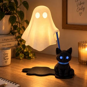 Op de afbeelding: Een tafellamp met een witte, gloeiende spookfiguur die boven een zwart kattenbeeldje zweeft. De kat heeft helderblauwe ogen en een blauwe halsband en houdt een blauwe staaf vast die het spook ondersteunt. De voet is een platte, zwarte, onregelmatige vorm.