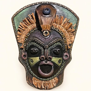 Könnte beinhalten: Eine dekorative Wandmaske mit einem dunklen Gesicht, markanten Augen, einer breiten Nase und einem offenen Mund. Die Maske ist mit strukturierten braunen und grünen Elementen, einer Franse aus hellbraunen Fasern und einem gemusterten Rand verziert.