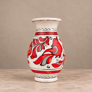 Puede incluir: Un jarrón de cerámica blanca con un motivo de pájaro rojo pintado a mano rodeado de patrones florales negros y rojos. El jarrón tiene un borde acampanado, un cuerpo redondeado y una base de pedestal, con bandas horizontales rojas y negras.