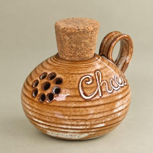 May include: A round, brown ceramic container with a cork stopper and a side handle. The surface features horizontal ridges, a cluster of seven circular holes on the left side, and the word Cheese written in white cursive script on the right side.