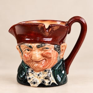 May include: A ceramic character jug shaped like a person wearing a brown tricorn hat, a dark green coat, and a white polka-dot cravat. The figure has a detailed facial expression with a prominent nose and a handle attached to the right side.