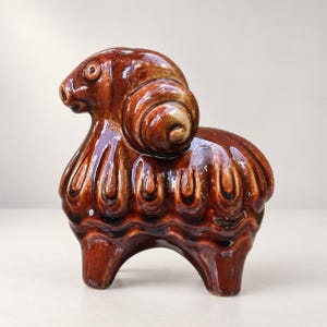 Könnte beinhalten: Eine glänzend braune Keramikfigur eines Widders mit einem stilisierten, eingerollten Horn an der Seite des Kopfes und einem strukturierten, wellenförmigen Muster entlang des Körpers. Die Skulptur hat eine glatte, reflektierende Oberfläche.