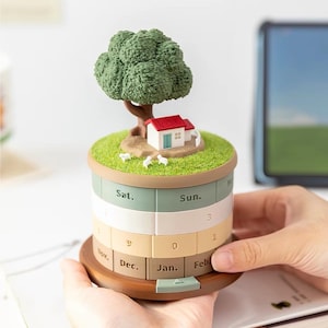 Puede incluir: Un calendario perpetuo cilíndrico que presenta un paisaje en miniatura en la parte superior con un árbol verde, una pequeña casa blanca con techo rojo y diminutas ovejas blancas sobre una base de césped. Los lados muestran paneles giratorios.