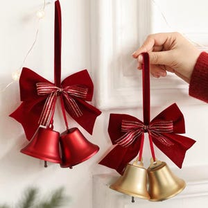 May include: Two hanging ornaments featuring pairs of bells attached to velvet ribbons and bows. One ornament has matte red bells, while the other has matte gold bells. Each bow is accented with a thin, striped ribbon detail in red and gold.