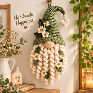 May include: A crocheted gnome wall hanging featuring a pointed olive green hat, a cream-coloured curly beard, and small white flowers with green leaves. A heart-shaped charm hangs from the hat tip. The text Handmade Happiness is visible.