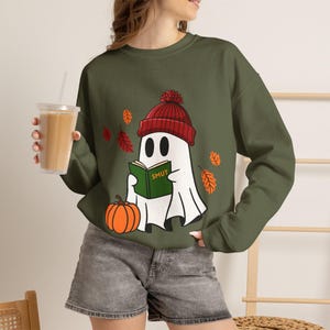 May include: An olive green crewneck sweatshirt featuring a graphic of a white ghost wearing a red knit beanie, reading a green book labeled SMUT, and standing next to an orange pumpkin with falling autumn leaves in red and orange colors.