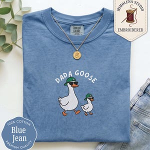 May include: A blue crew neck t-shirt featuring an embroidered graphic of a large goose and a small gosling, both wearing green baseball caps and black sunglasses. The text DADA GOOSE is embroidered in white above the geese. The shirt has a Comfort Colors label at the inner neckline.