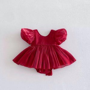 May include: A deep red baby romper featuring short, voluminous puff sleeves and a wide, rounded neckline. The bodice is fitted, transitioning into a full, pleated skirt-like overlay made of a sheer, lightweight fabric with a subtle sheen.