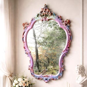 Puede incluir: Espejo ornamentado con un marco multicolor en tonos rosa, morado y azul. El espejo refleja una escena natural al aire libre. Está montado en una pared blanca sobre una mesa de mármol blanco.