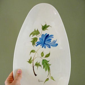 Puede incluir: Plato ovalado de cerámica blanquecina con una flor azul y hojas verdes pintadas a mano. El diseño floral está centrado en el plato. El plato es sostenido por una mano.