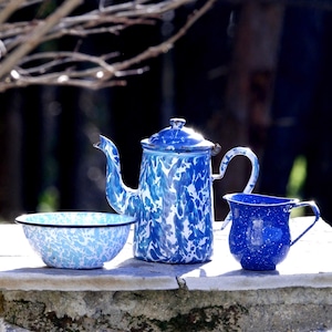 Könnte beinhalten: Eine Sammlung von Küchenartikeln aus Emaille im Vintage-Stil, darunter eine Teekanne, ein kleiner Krug und eine Schüssel. Die Artikel weisen ein blau-weißes Sprenkelmuster auf. Die Teekanne hat einen Deckel und einen gebogenen Ausguss. Krug und Schüssel sind kleiner und haben ein ähnliches Design.