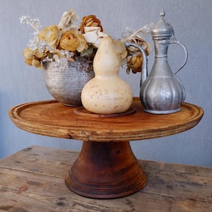 May include: A rustic wooden cake stand with a round top, holding a decorative gourd, a silver metal pitcher, and a small metal bucket filled with dried flowers. The stand is made of dark brown wood.