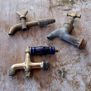 May include: Four antique brass taps are displayed on a distressed wooden surface. The taps have different designs, including a blue-handled tap and others with cross or lever handles. The brass shows signs of age and wear, with a tarnished finish.