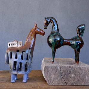 Puede incluir: Dos esculturas de caballos de cerámica. Una es marrón y azul con una estructura similar a un edificio en su espalda. La otra es verde oscuro y roja, con una forma estilizada única. Ambas están sobre una superficie de madera sobre un fondo gris.