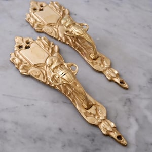 Puede incluir: Dos tiradores de cajón dorados y ornamentados con un diseño clásico. Cada tirador presenta un elemento en forma de escudo, una manija central y detalles florales y de volutas decorativas. Los tiradores son probablemente de metal.