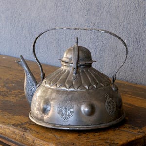 May include: A silver metal teapot with a curved handle and a decorative spout. The teapot has embossed floral patterns and a hinged lid. It is displayed on a wooden surface.