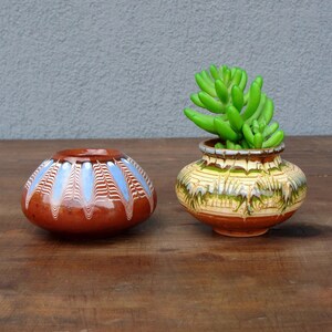 May include: Two ceramic pots on a wooden surface. One is brown with blue and white patterns, the other is yellow, green, and brown, holding a green succulent plant. The pots have a rustic, handmade appearance.