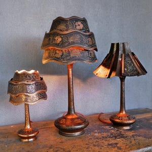 May include: Three antique copper lamps with detailed floral patterns. The lampshades have a tiered, scalloped edge. The bases and stems are etched. The lamps are lit, emitting a warm glow. The lamps are on a wooden surface.