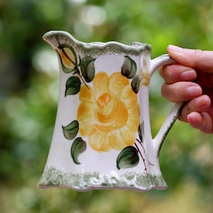 Op de afbeelding: Witte keramische kan met een gele bloem en groene bladeren. De kan heeft een geschulpte rand en een handvat. Het bloemmotief is gecentreerd op de voorkant. De kan wordt door een hand vastgehouden.