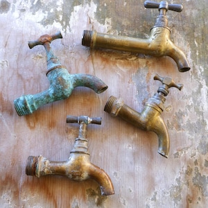 May include: Four antique brass taps with a weathered appearance are arranged on a wooden surface. The taps have a tarnished, aged look, with some showing green oxidation. The handles are cross-shaped.