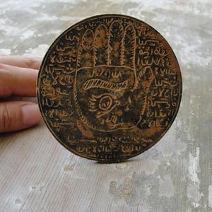 Op de afbeelding: Een ronde bronzen medaillon in antieke stijl. Het toont een gedetailleerd handsymbool met een oog in de palm, omringd door ingewikkelde tekst. Het medaillon heeft een verweerde, verouderde uitstraling.