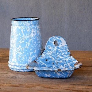Könnte beinhalten: Ein blau-weißes Emaille-Set, bestehend aus einem hohen zylindrischen Behälter und einer Seifenschale mit Deckel. Die Teile haben ein gesprenkeltes Muster und einen schwarzen Rand. Das Set wird auf einer Holzoberfläche präsentiert.