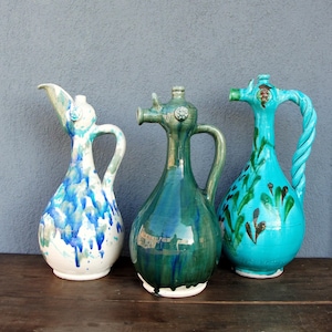 May include: Three ceramic jugs with distinctive designs. The jug on the left is white with blue paint drips. The middle jug is green, and the right jug is turquoise with a twisted handle and green and brown accents.